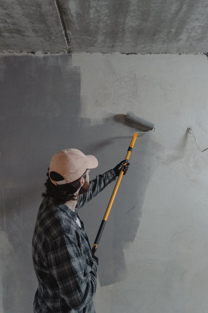 A man using a paint roller to apply gray paint on a wall in an unfinished room.
